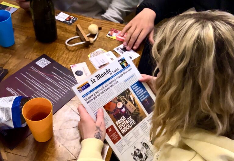 Joueuse tenant un journal "Le Monde de l'apéro" avec un article sur une prise d'otage insolite, jeu "Escape Games L'Apéro"